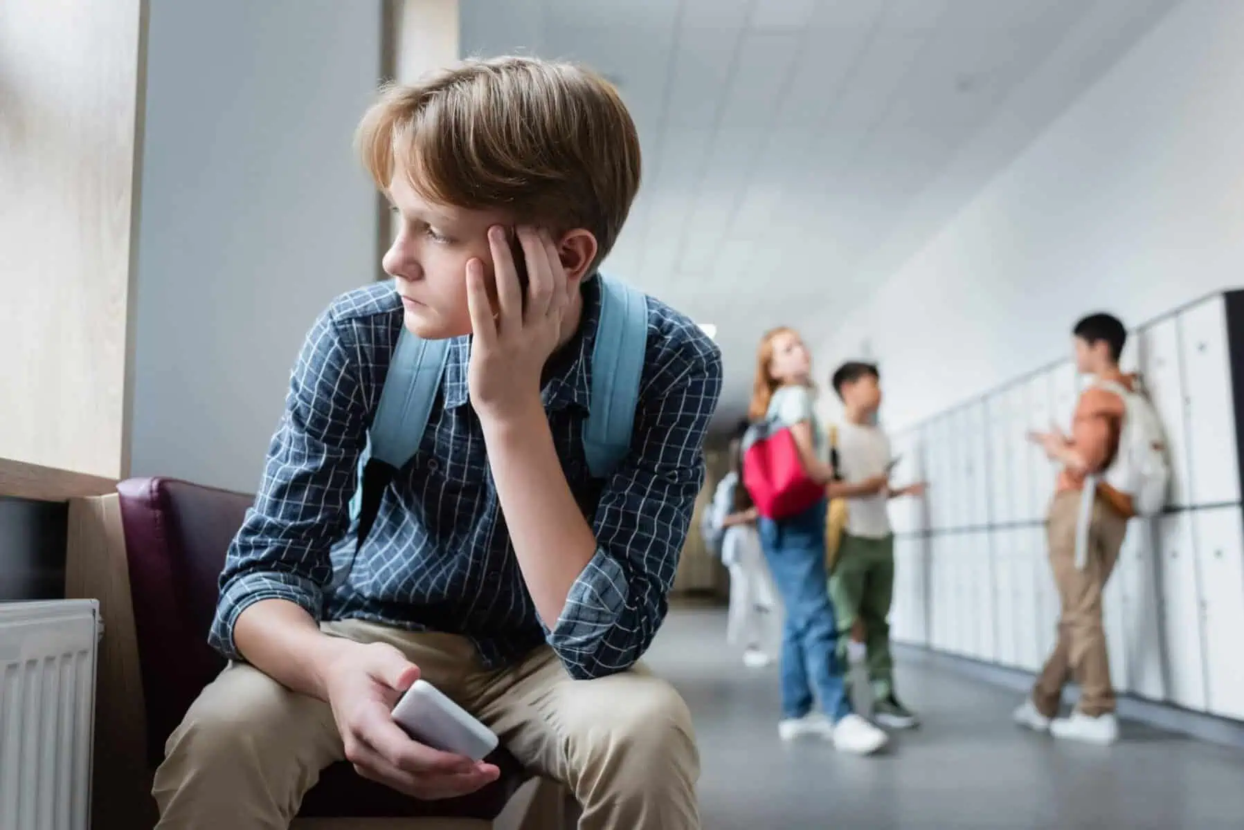 depressed boy with smartphone sitting alone in school corridor near teenagers on blurred background