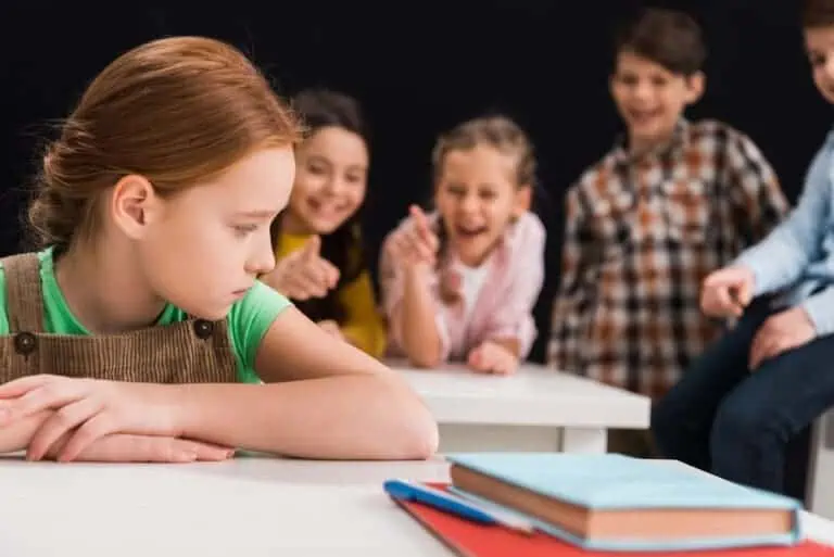 selective focus of upset schoolkid sitting near classmates laughing and pointing with fingers isolated on black, bullying concept