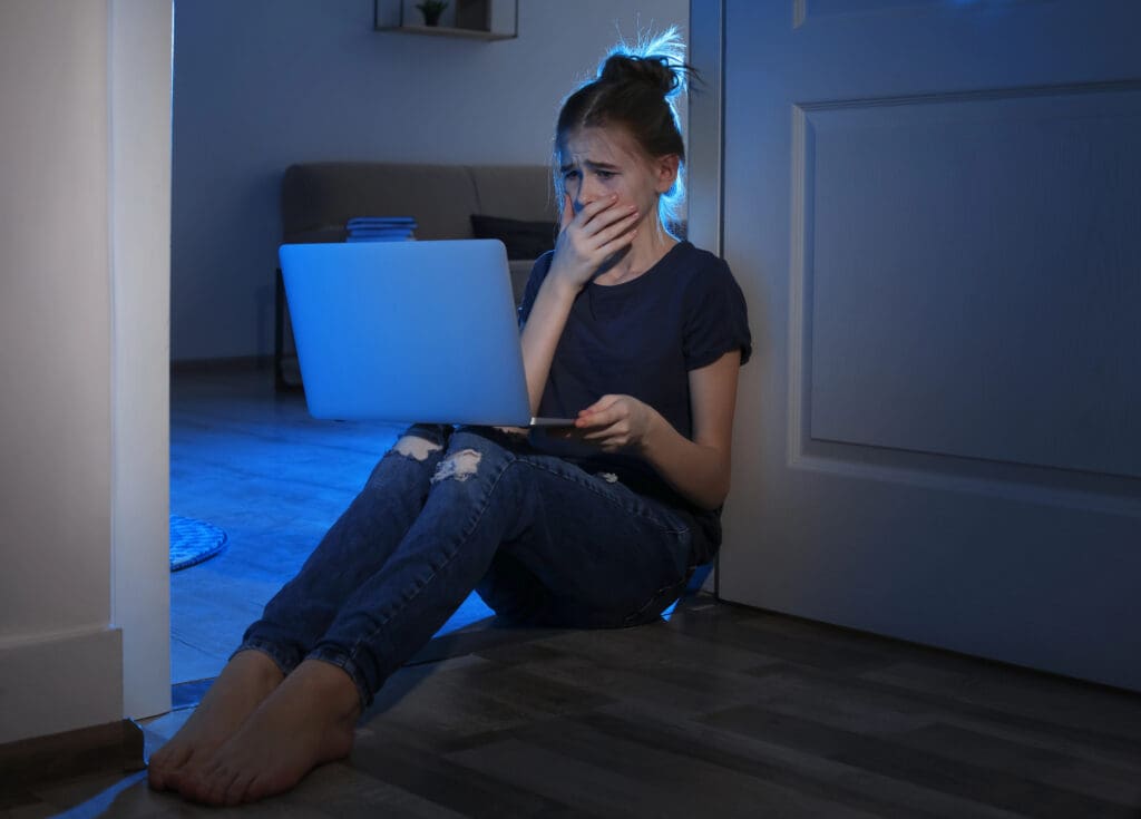 Frightened teenage girl with laptop on floor in dark room.