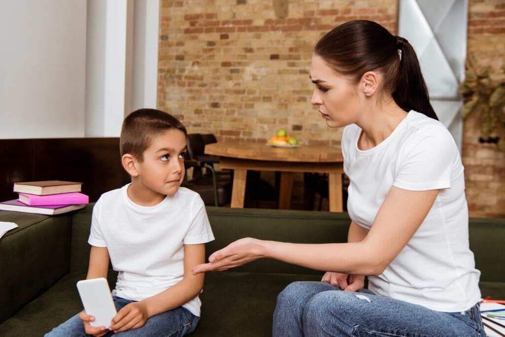 Angry mother pointing with hand at son holding smartphone on sofa