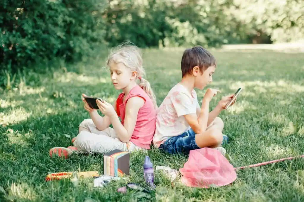 Boy and girl play games on smartphones outdoor. Kids digital gadget screen addiction. Children friends playing online sitting on grass in park. Contemporary problem of loneliness together.