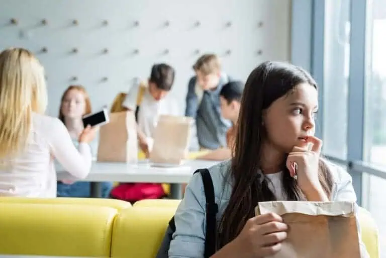 depressed schoolgirl looking away while sitting with paper bag in canteen near blurred teenagers