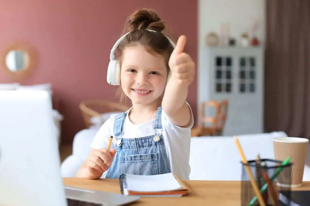 Happy little girl pupil study online using laptop at home, smiling small child show thumb up recommend class or lesson