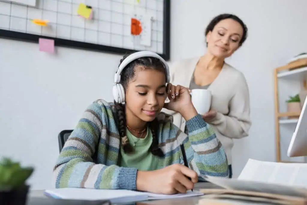 african american girl in headphones writing in notebook near blurred mom with cup of tea
