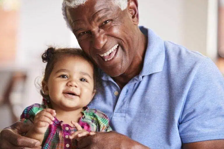 Portrait Of Smiling Grandfather Sitting On Sofa At Home With Baby Granddaughter