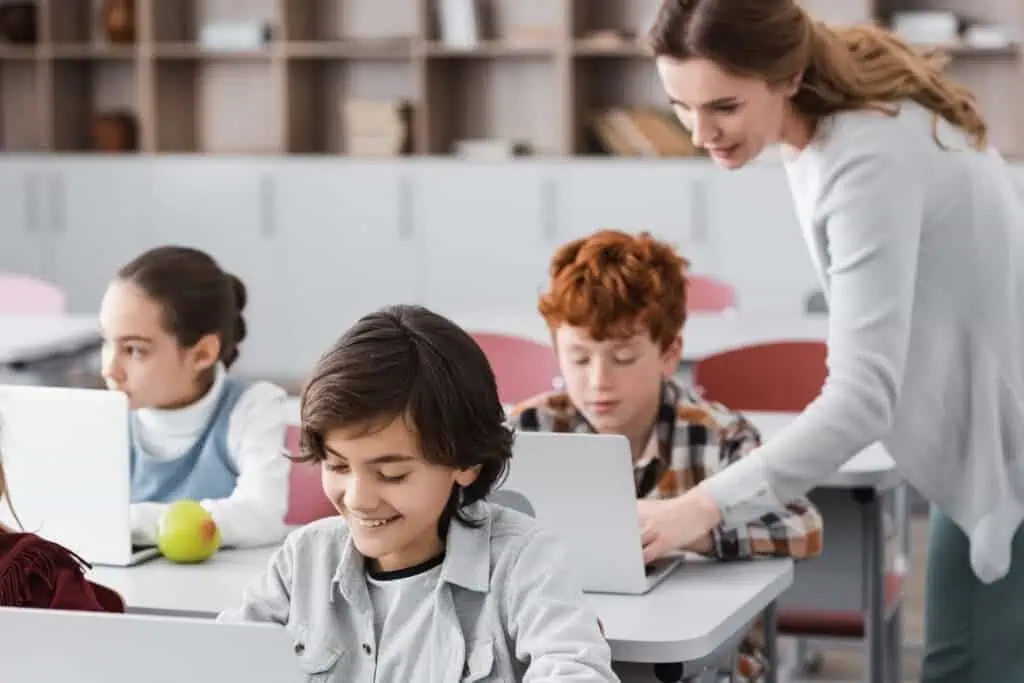 teacher helping schoolboy working on laptop during lesson, blurred background