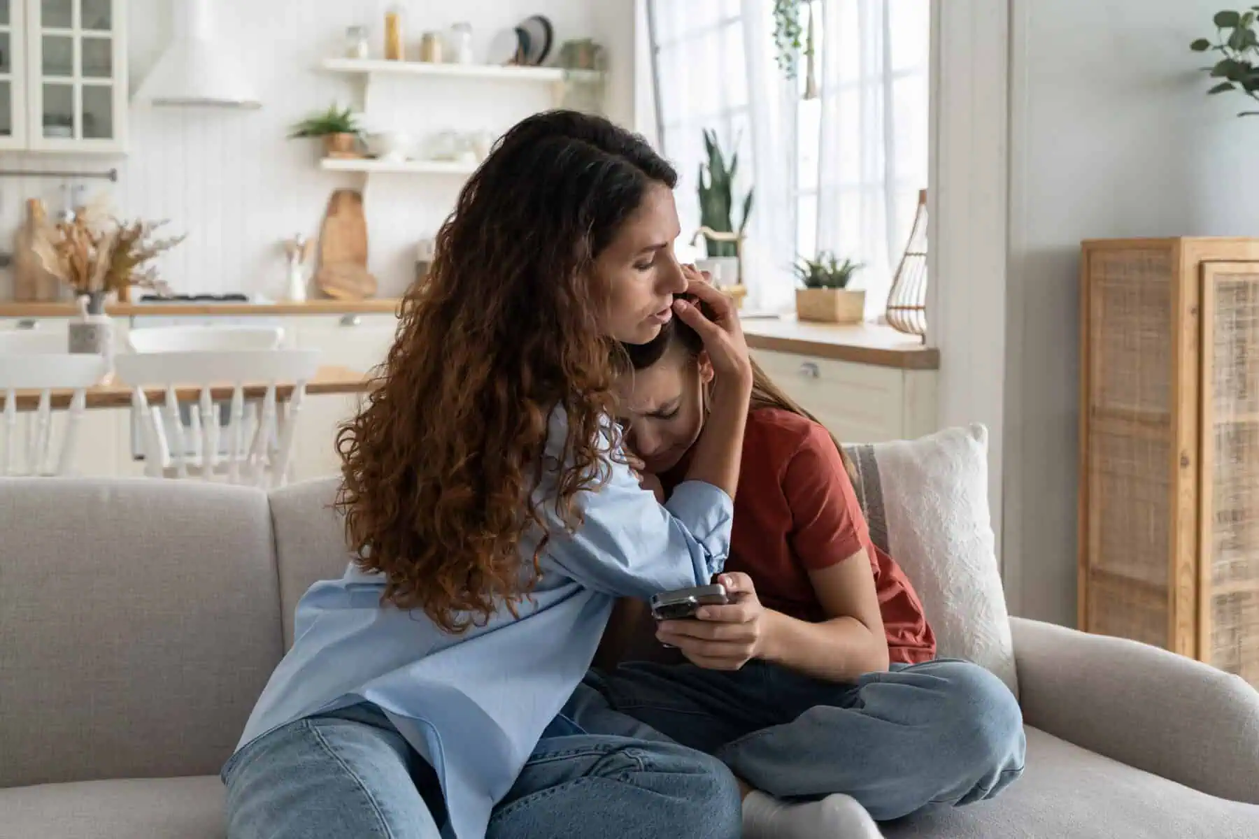 Upset crying teenage girl sits on sofa with phone suffering from depression after bullying. Caring kind woman hugging daughter feeling stressed because of insults in social networks or messengers
