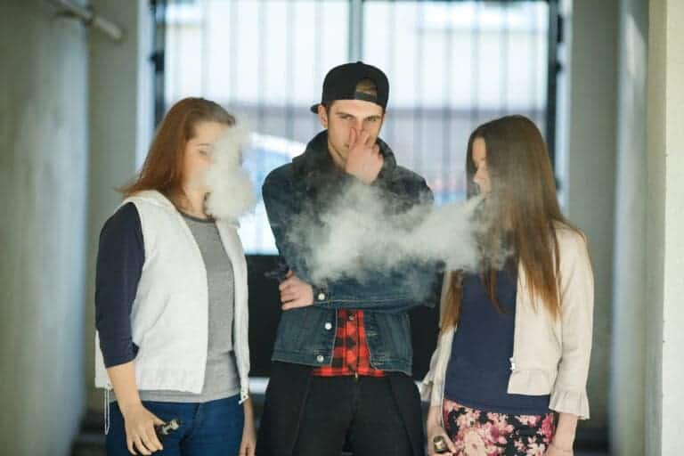 Vape teenagers. Two young cute girls in casual clothes smoke electronic cigarettes near nonsmoking guy outdoors in the street in summer day.