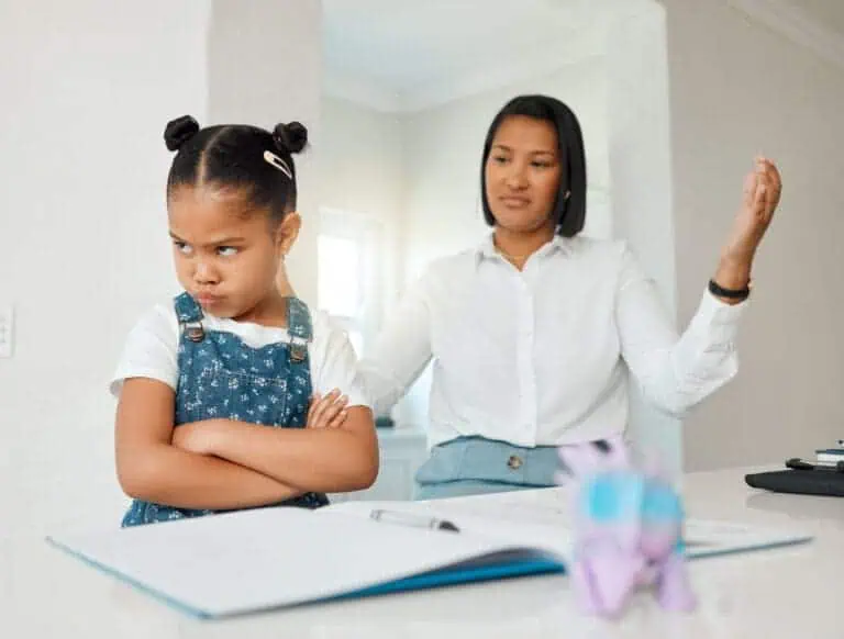 Girl is frustrated at table with notebook and crayon while mom talks in background