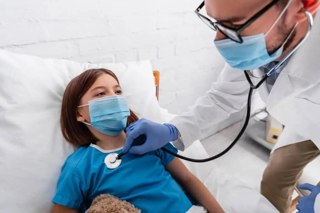 pediatrician in medical mask examining sick girl with stethoscope in hospital