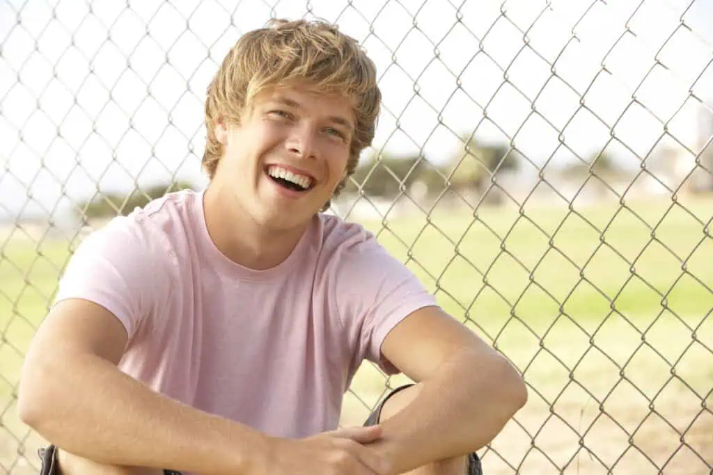 Teen boy smiling, sitting outside near fence. 