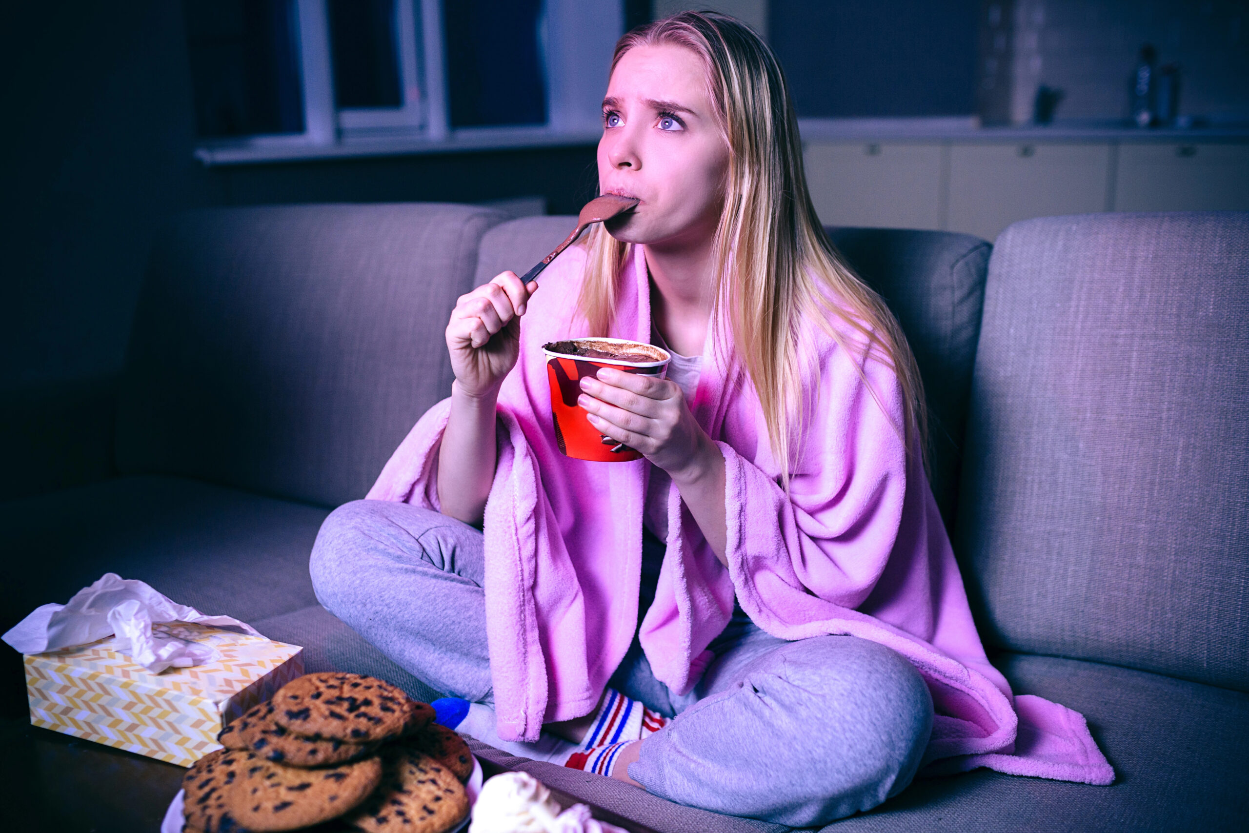 Young woman watching movie at night. Concentrated model eating ice cream with spoon. Sitting on sofa in room alone. Streaming show or tv series.