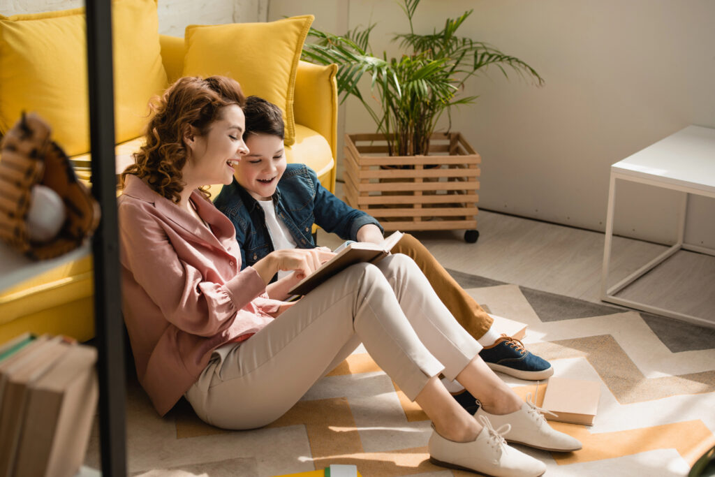 A mom sits in the floor enjoying a book with her delighted son.