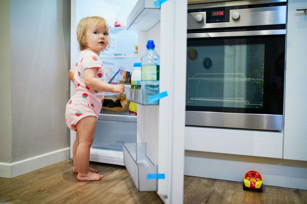 Adorable toddler girl at home, opening the fridge and selecting food