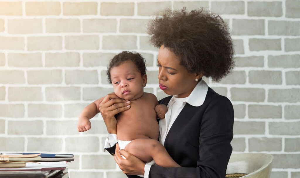 African mother in business suit holding son while working at home. Uncomfortable infant with stressful mom. Business, single mom or Women's day concept.