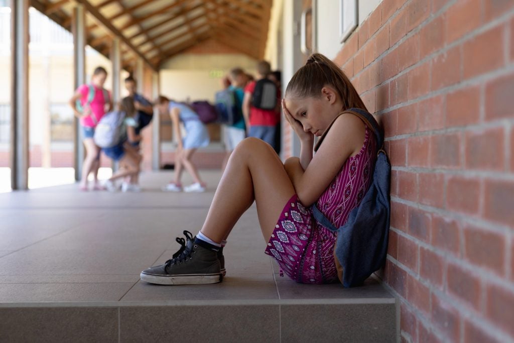 Side view of a Caucasian schoolgirl wearing a purple dress and a rucksack sitting on the ground against a wall alone in the schoolyard at elementary school looking sad, with other schoolchildren standing together in the background