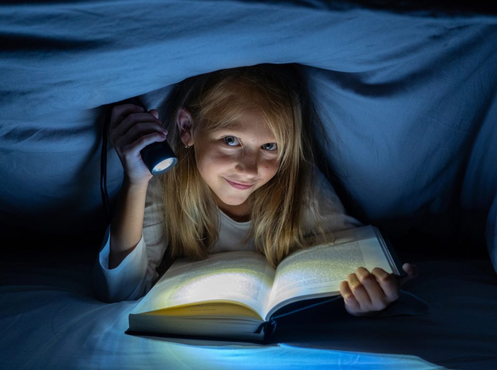 Happy smart girl reading a fairytale book lying in the dark under the cover bed holding a lantern in Reading skills Literature School Success and Education concept.