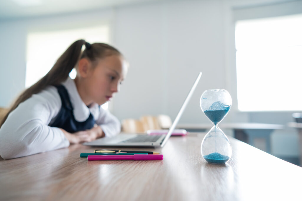 Caucasian girl sits at a desk at school and studies at a laptop. Hourglass on the table