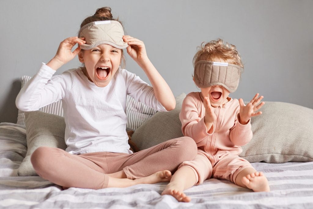 Portrait of crazy funny little girls playing in bedroom, sisters with blindfold waking up in good playful mood sitting on bed and screaming yelling with happiness.