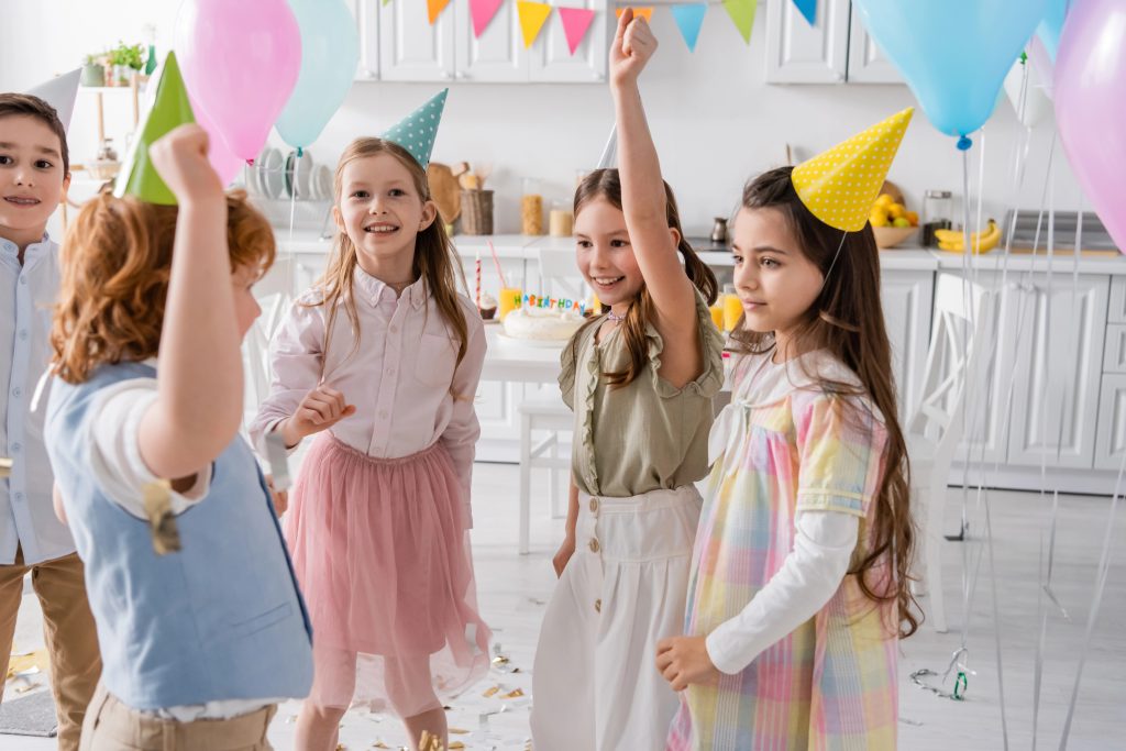 group of happy children in party caps dancing during birthday celebration at home