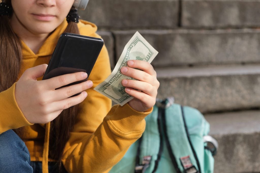 The teenager holds dollar bills and looks at the smartphone screen.