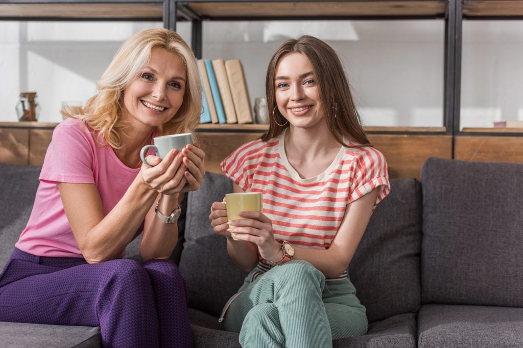 cheerful mother and daughter looking at camera while sitting on sofa and holding tea cups