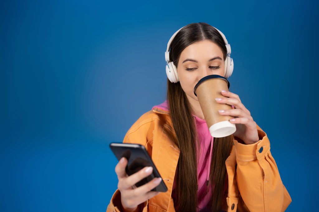 Teenage girl in headphones chatting on smartphone and drinking coffee to go isolated on blue