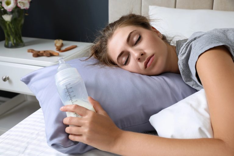 Exhausted young mother postpartum with bottle of milk sleeping in bed at home