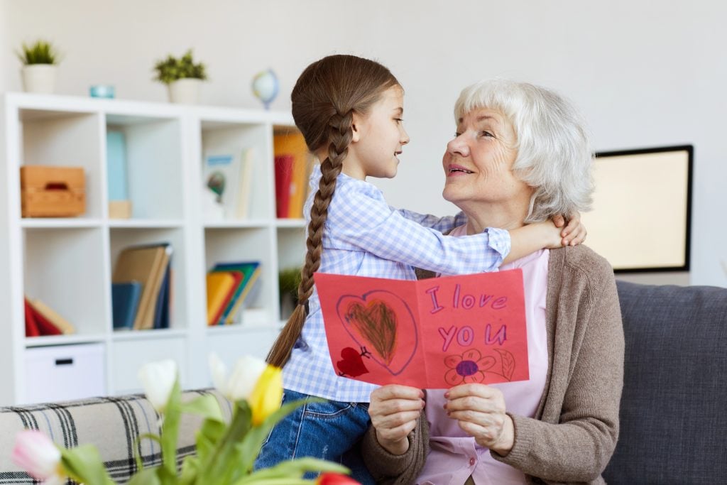 Portrait of cute little girl hugging grandma and giving her handmade card on Birthday, copy space