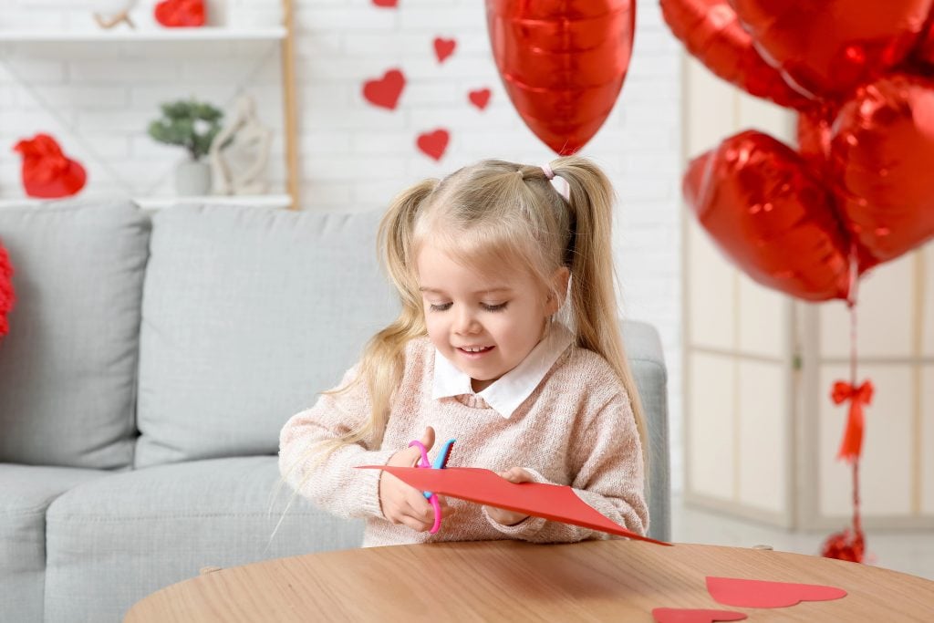 Cute little girl cutting paper hearts in room decorated for Valentines Day