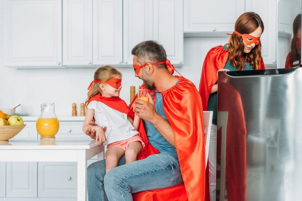 family in costumes of superheroes spending time in kitchen, while father and daughter drinking orange juice and mother standing near fridge