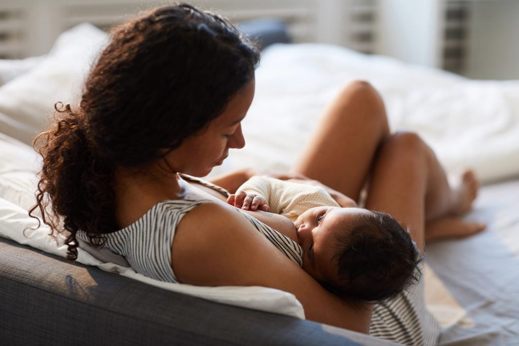 Peaceful loving young African mother sitting on bed and leaning on headboard while feeding baby with breast