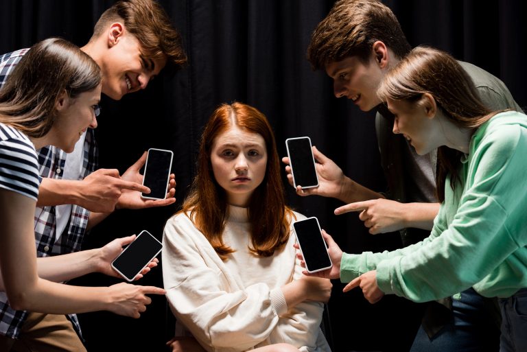 smiling teenagers pointing with fingers at girl during bullying and holding smartphones with blank screen on black