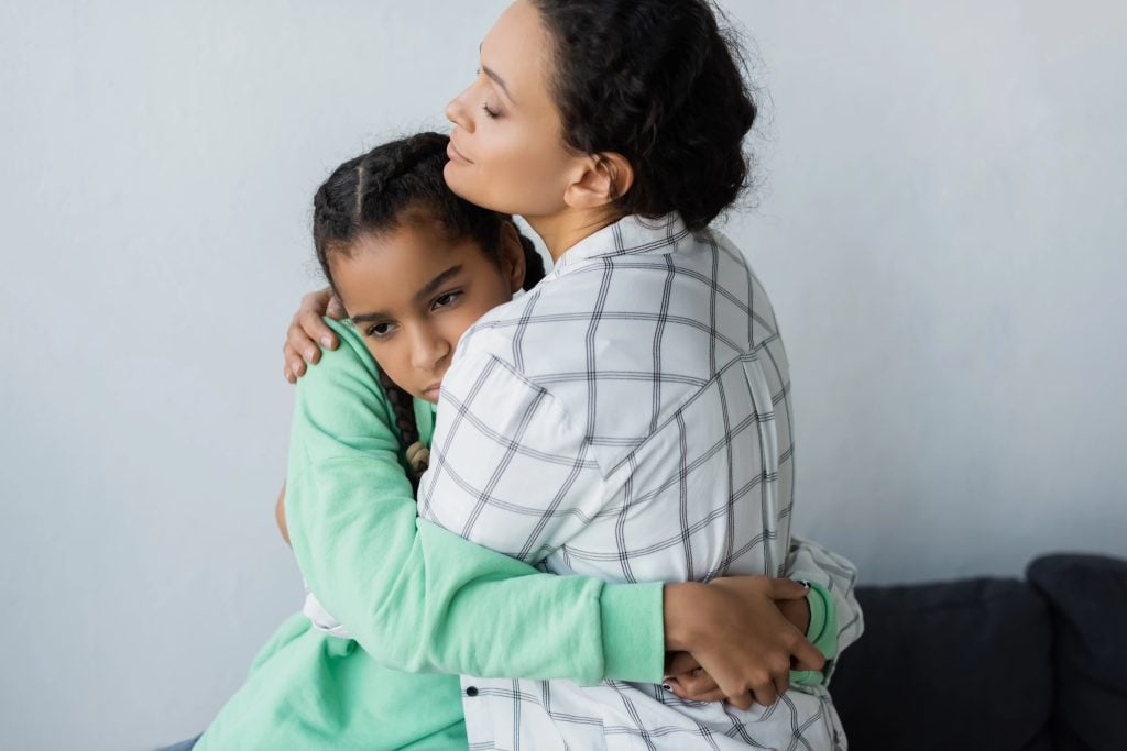 african american mother with closed eyes embracing depressed teenage daughter at home