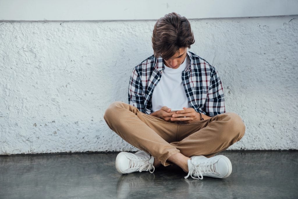 front view of teenager sitting with crossed legs and using smartphone