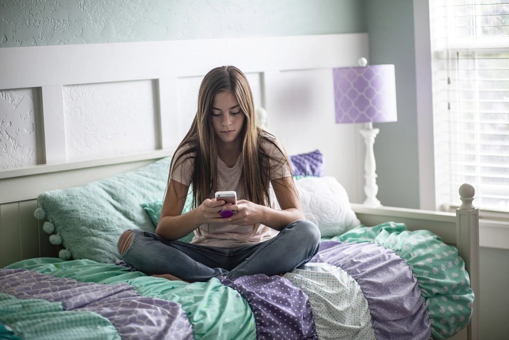 Adolescent teen girl texting on a smartphone sitting in bed at home in her bedroom