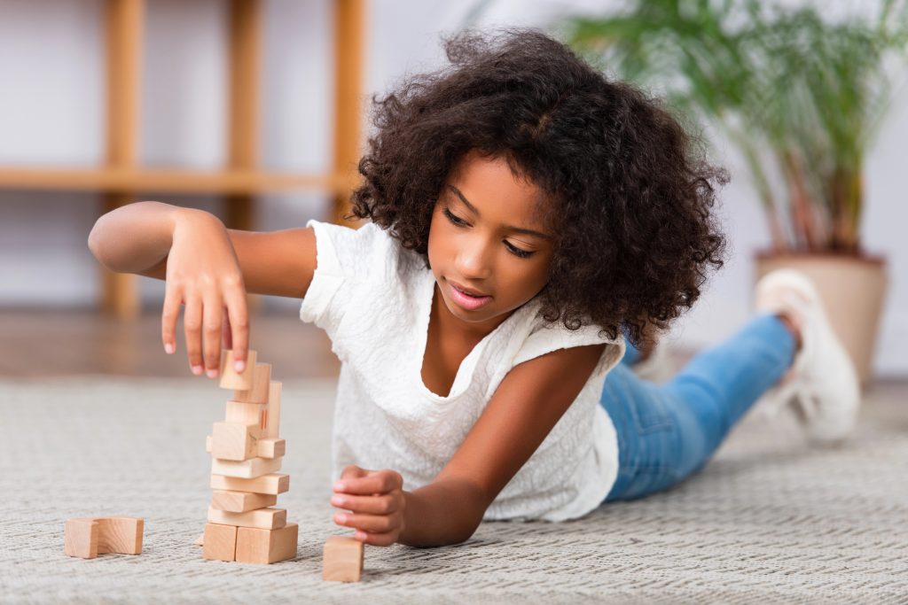 Concentrated african american girl playing with wooden blocks while lying on floor with blurred office on background
