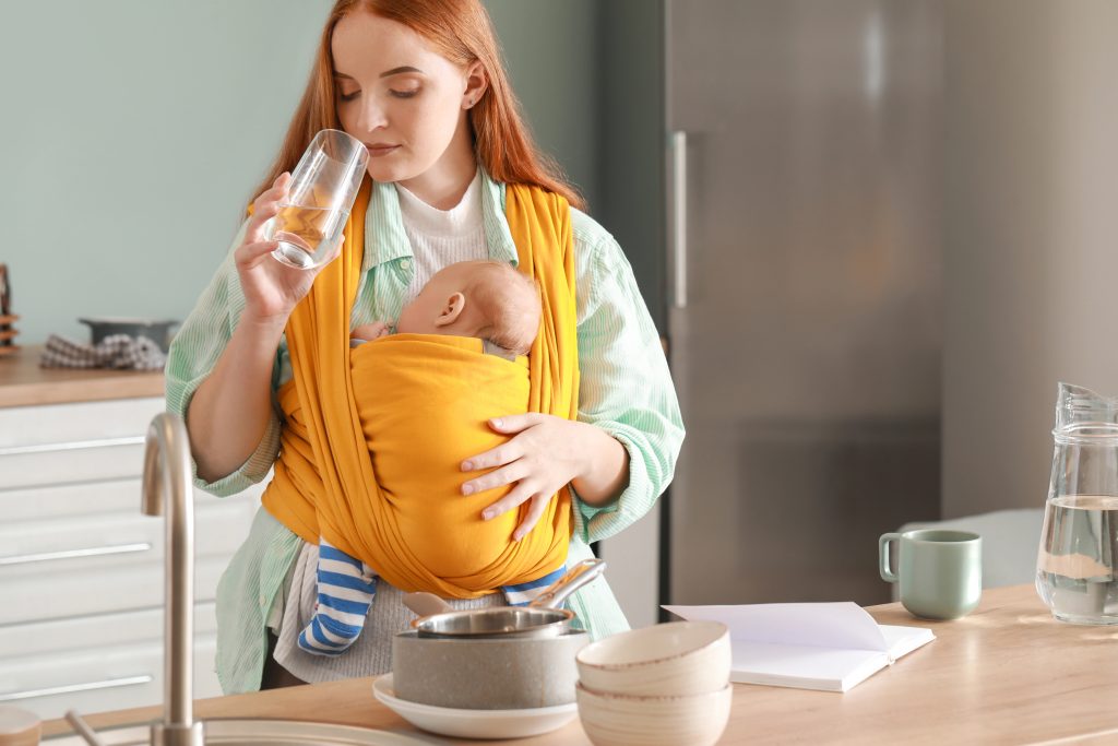 Young mother with little baby in sling drinking water at home