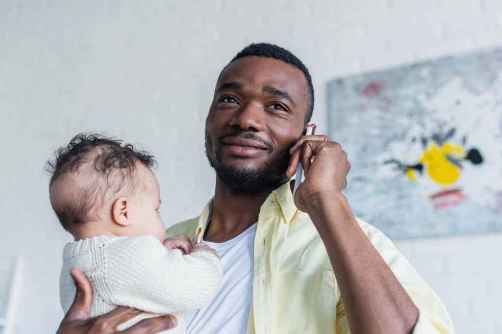 positive african american man holding infant kid while talking on mobile phone
