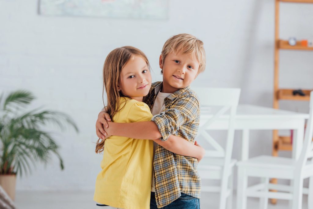 brother and sister embracing and looking at camera while standing at home