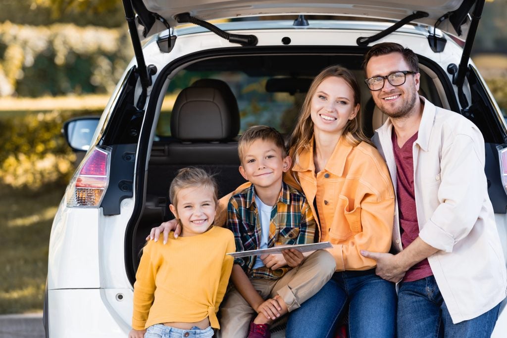 Cheerful family with map looking at camera near trunk of car outdoors