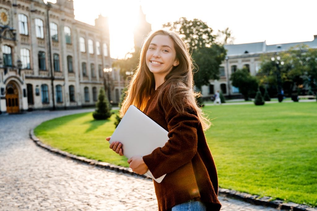 Charming young woman student in move walking in university garden with laptop computer, going to her classroom, beautiful sunset background, positive emotions, lifestyle people
