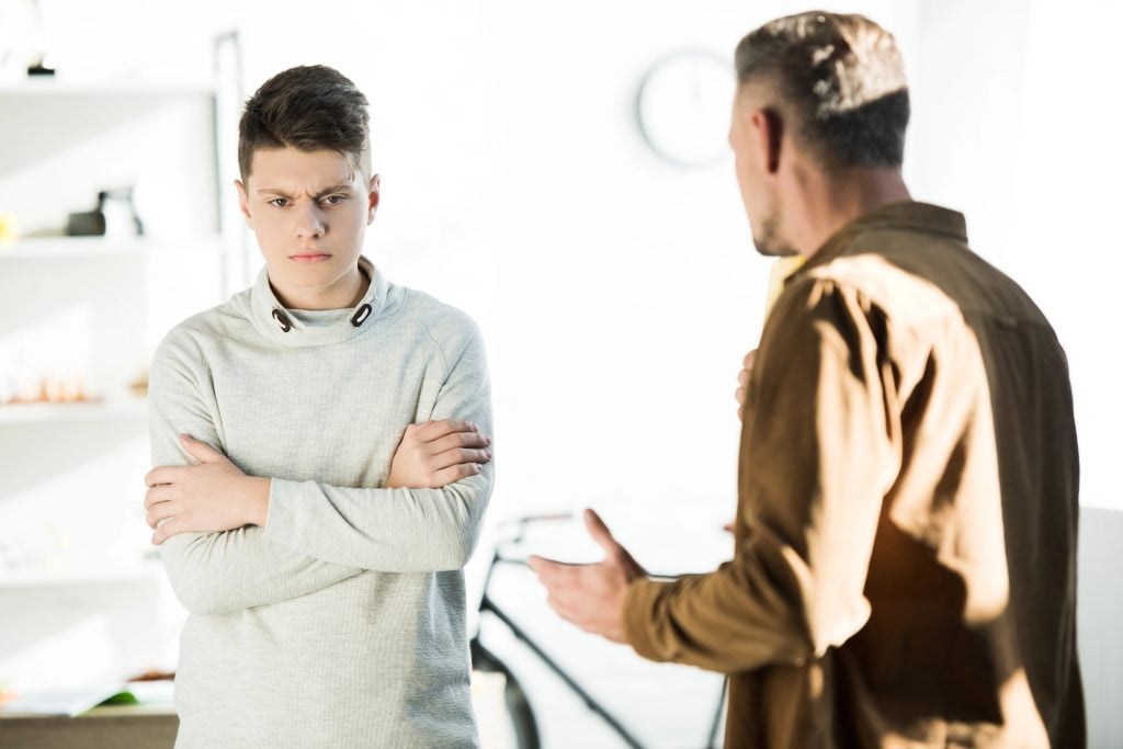 irritated father gesturing and looking at teen son with crossed arms at home