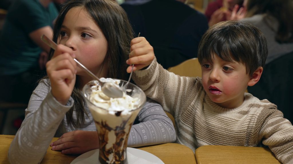 Kdis eating whipped cream sundae at restaurant, brother and sister sharing sugar treat indulgence at diner in the evening weekend outing