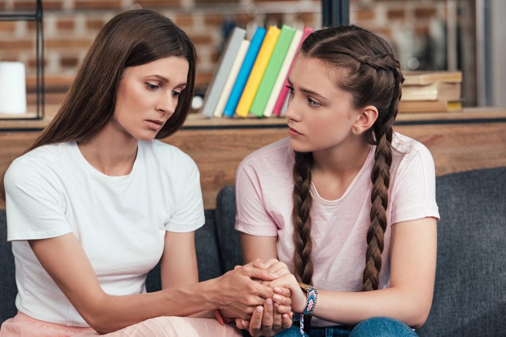 selective focus of teenage girl holding hands of frustrated mother on sofa