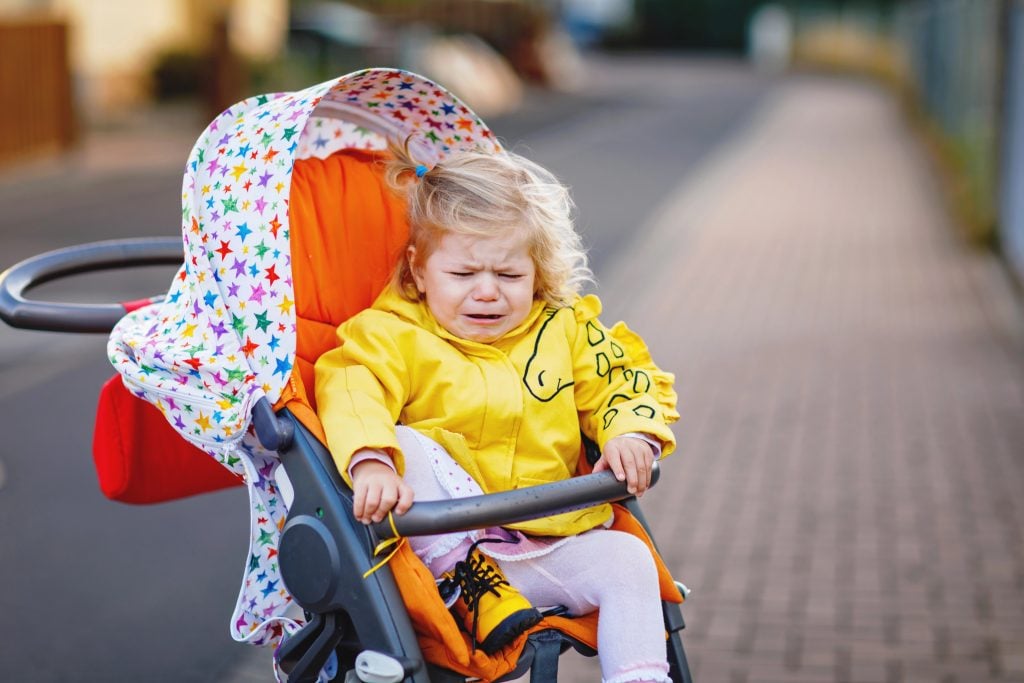 Portrait of little sad toddler girl sitting in stroller and going for a walk. Crying baby child does not want sitting in pram. Healthy daughter. Hysteric crisis of two years phase.