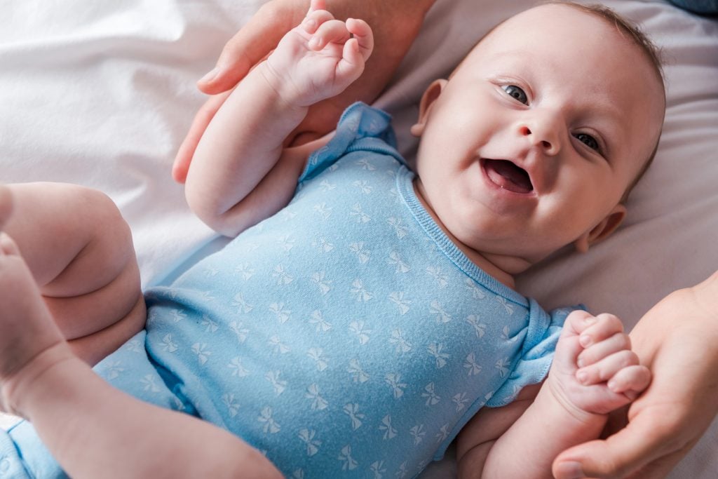 cropped view of woman holding hands of happy infant in blue baby bodysuit