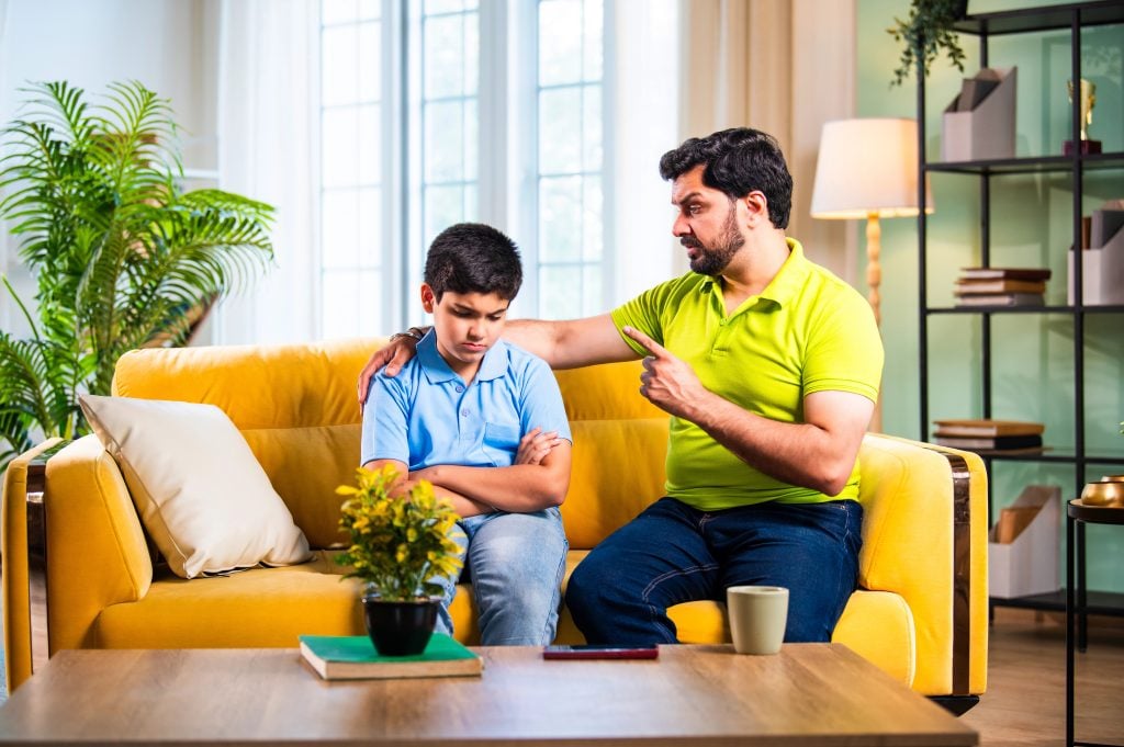 Indian asian young father scolding son while sitting on sofa in modern home as upset child turns back silently, showing emotional parenting moment and family tension inside living room