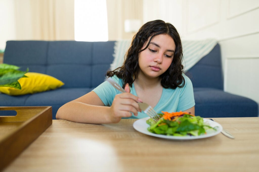 Unwilling teenage girl eating a healthy salad at home, grappling with eating disorder and health issues, highlighting the connection between diet and mental health