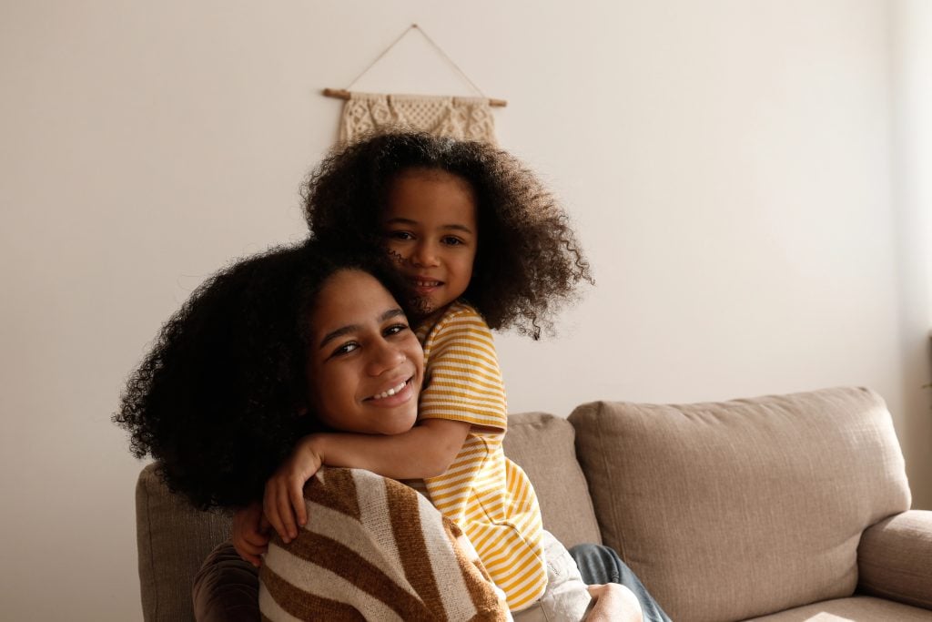 Younger and older sister spending time together at home. Two black girls of different age hugging and showing affection. Black female siblings having fun and bonding. Background, copy space, close up.